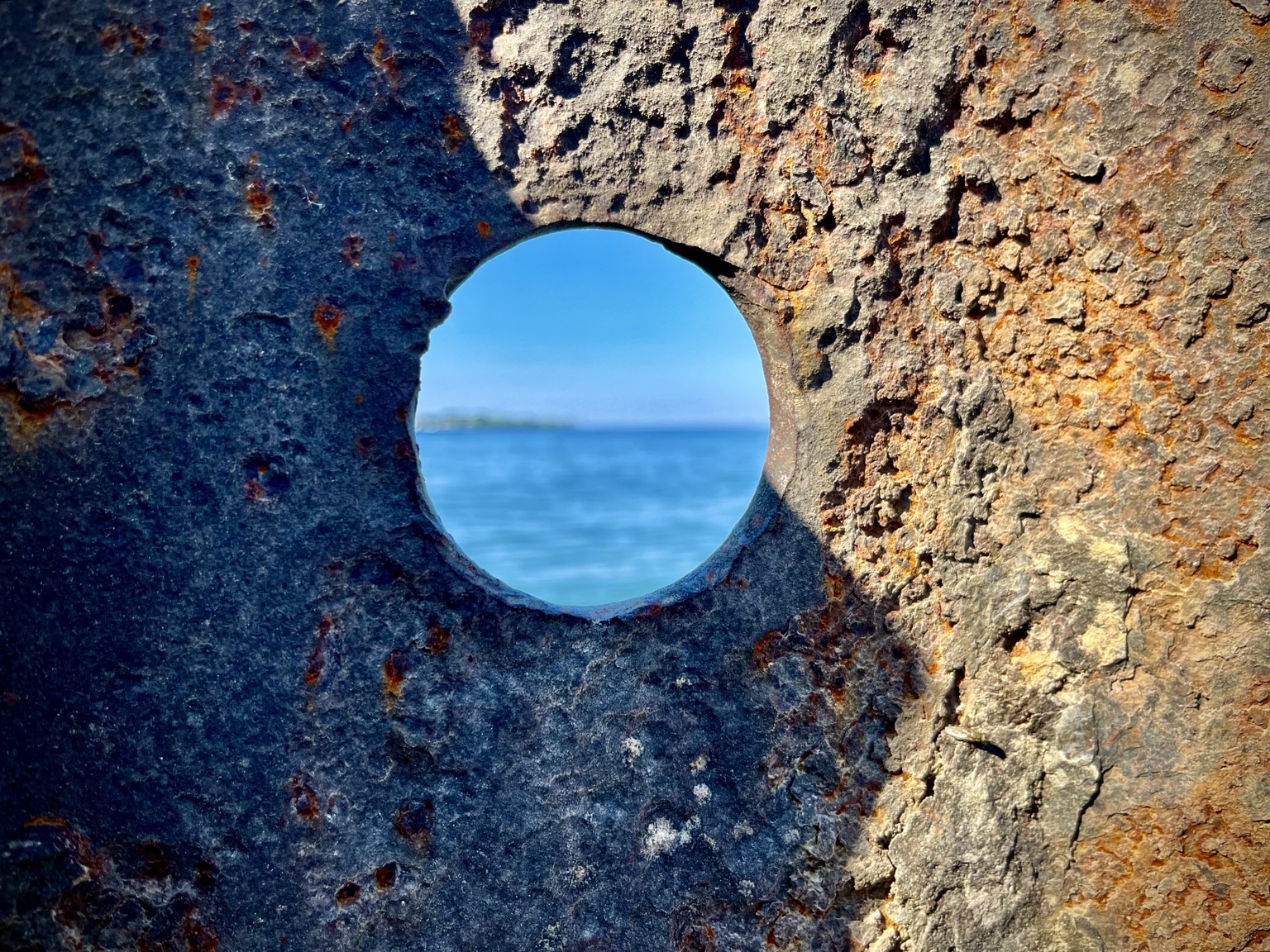 Blue water seen through a circular opening in weathered metal.